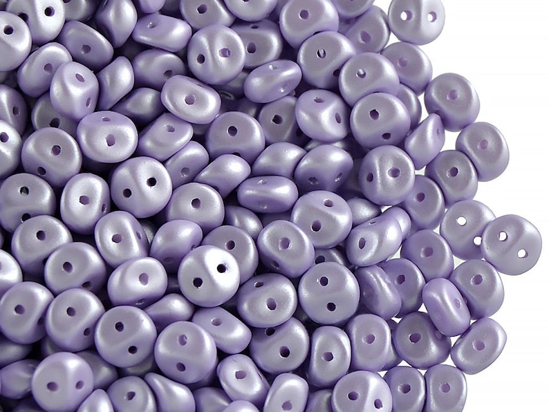 May include: A pile of small, light purple, two-hole beads. The beads are smooth and have a pearlescent finish.