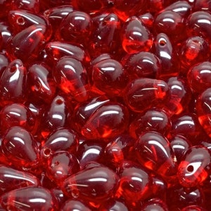 May include: A collection of numerous small, translucent, ruby red glass beads shaped like teardrops. Each bead features a smooth, polished surface with a reflective sheen and a single small hole drilled near the top for threading purposes.