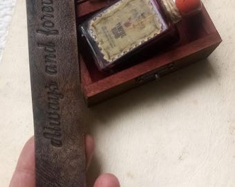 May include: A small wooden box with a hinged lid, containing a glass bottle with a red liquid and a label that reads "Vampire Blood". The box is open and the bottle is visible. A brown leather bookmark with the text "Always and Forever" is in the foreground.