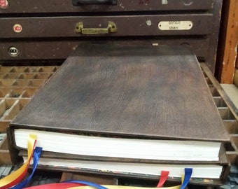 May include: Two brown leather-bound books with white pages are stacked on top of each other. The books are tied together with three ribbons: red, blue, and yellow. The books are sitting on a wooden surface.