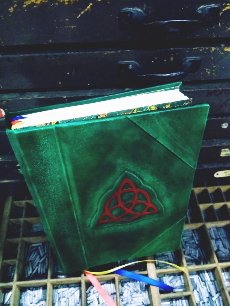 May include: A green leather-bound journal with a red, triangular symbol on the front cover. The journal is sitting on a wooden surface with a grid of compartments.