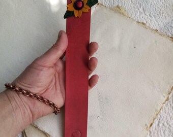 May include: A red bookmark with a sunflower design at the top. The sunflower has yellow petals, a purple center, and green leaves. The bookmark is held in a hand.