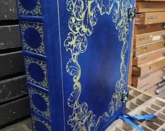 May include: A blue leather-bound book with gold decorative accents and a gold-colored border. The book is closed and has a ribbon bookmark.