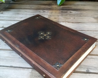 May include: A brown leather-bound journal with decorative brass accents in the shape of flowers. The journal has a white interior and is closed.