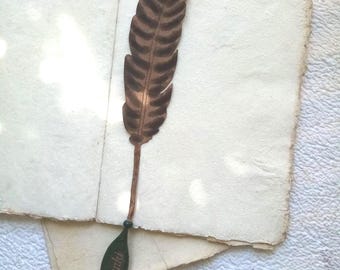 May include: A brown feather bookmark rests on the open pages of a vintage-style notebook. The bookmark has a dark brown feather design with a green leaf-shaped base that reads "Wanki". The notebook pages are off-white with rough edges.