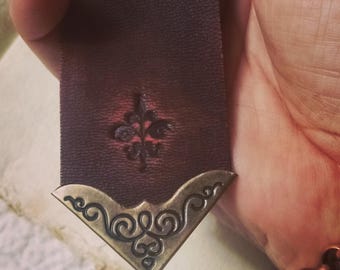May include: A dark brown leather bookmark with a decorative metal corner piece. The bookmark features a stamped floral design and a bronze-colored metal corner with an ornate scroll pattern. A unique accessory for book lovers.