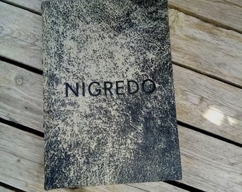 May include: A rectangular book with a textured, mottled surface in shades of gray and black. The word "NIGREDO" is printed in dark blue across the center of the cover. The book is resting on a wooden surface.