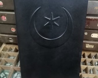 May include: Black leather-bound journal with a silver crescent moon and star design embossed on the front cover. The journal is closed and resting on a wooden surface.