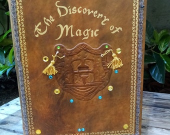 May include: A brown leather-bound book with gold lettering that reads "The Discovery of Magic" and decorative gold trim. The book has a gold tassel on each side and is adorned with gold and blue jewels.