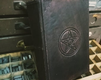 May include: A dark brown leather-bound book with a pentagram embossed on the front cover. The book is resting on a wooden tray filled with metal type.