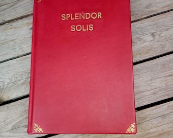May include: A red book with the title "SPLENDOR SOLIS" in gold lettering. The book has gold corner accents and is set against a wooden background. The cover appears to be leather or a similar material.