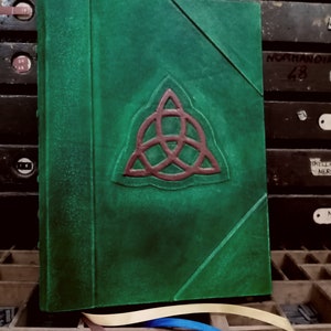 May include: A green leather-bound journal with a red Celtic knot design on the front cover. The journal is sitting on a wooden surface with a variety of metal type.