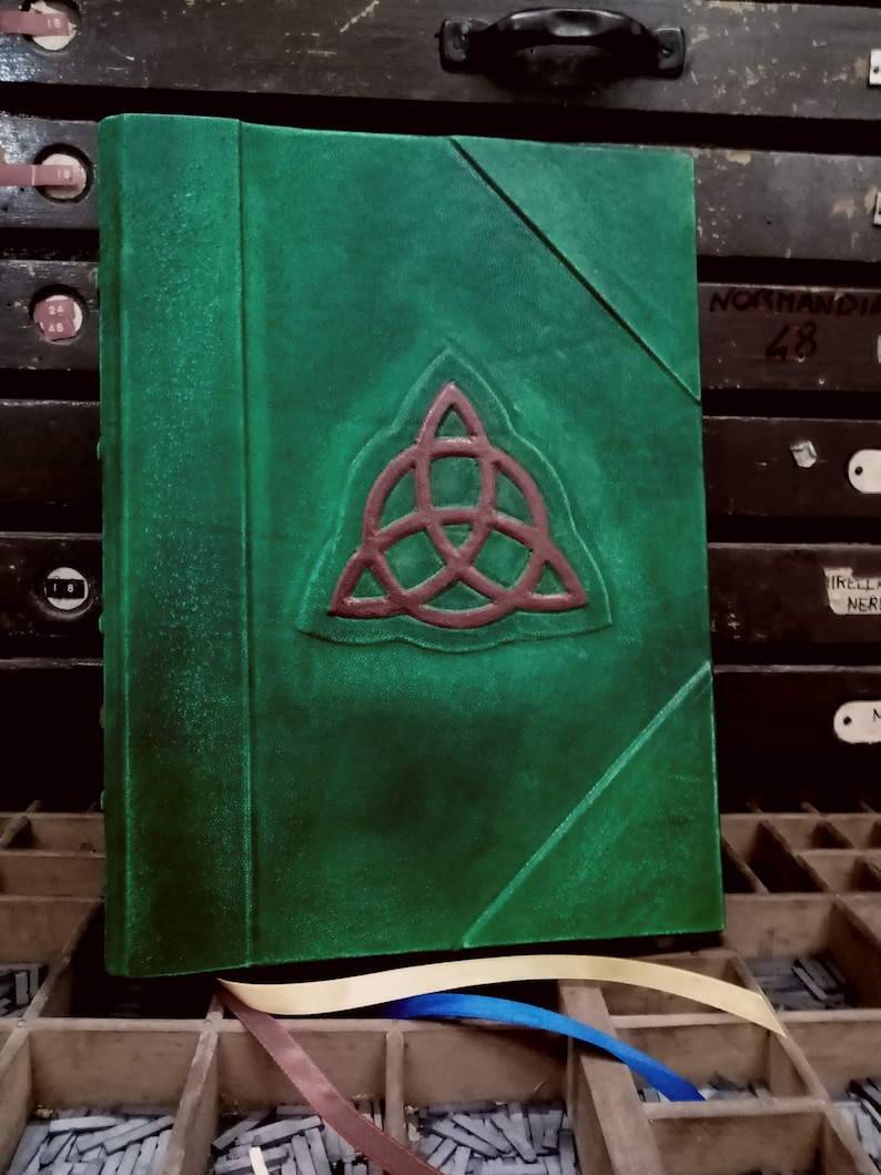 May include: A green leather-bound journal with a red Celtic knot design on the front cover. The journal is sitting on a wooden surface with a variety of metal type.