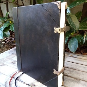 May include: A large, rectangular, black leather-bound book featuring decorative brass corner protectors and two brass metal clasps on the side. The book has thick, white paper pages and two thin ribbon bookmarks in red and black extending from the bottom.