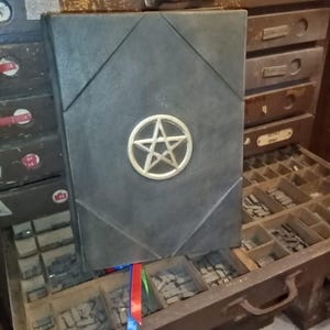 May include: A dark grey book featuring a gold pentagram emblem on the cover. The book has a geometric design and colourful ribbons marking the pages. The book is displayed in front of a wooden drawer.