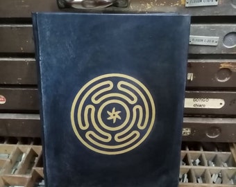 May include: A black leather-bound journal with a gold circular design featuring a star in the center. The journal is resting on a wooden surface with metal type.