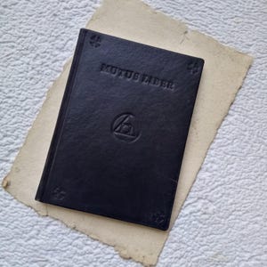 Op de afbeelding: Een zwart lederen gebonden boek met de titel "MUTUS LIBER" gegraveerd op de voorzijde. Een symbool van een cirkel met een driehoek erin is ook gegraveerd op de omslag.