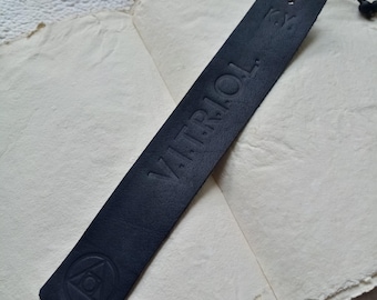 May include: Black leather bookmark with the word "VITRIOL" embossed in white lettering and a small triangle symbol with a circle inside. The bookmark has a black string attached to it.