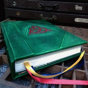 May include: A green leather-bound journal with a red and green Celtic knot design on the cover. The journal is closed and has a white ribbon bookmark. The journal is sitting on a wooden surface with a variety of metal type.
