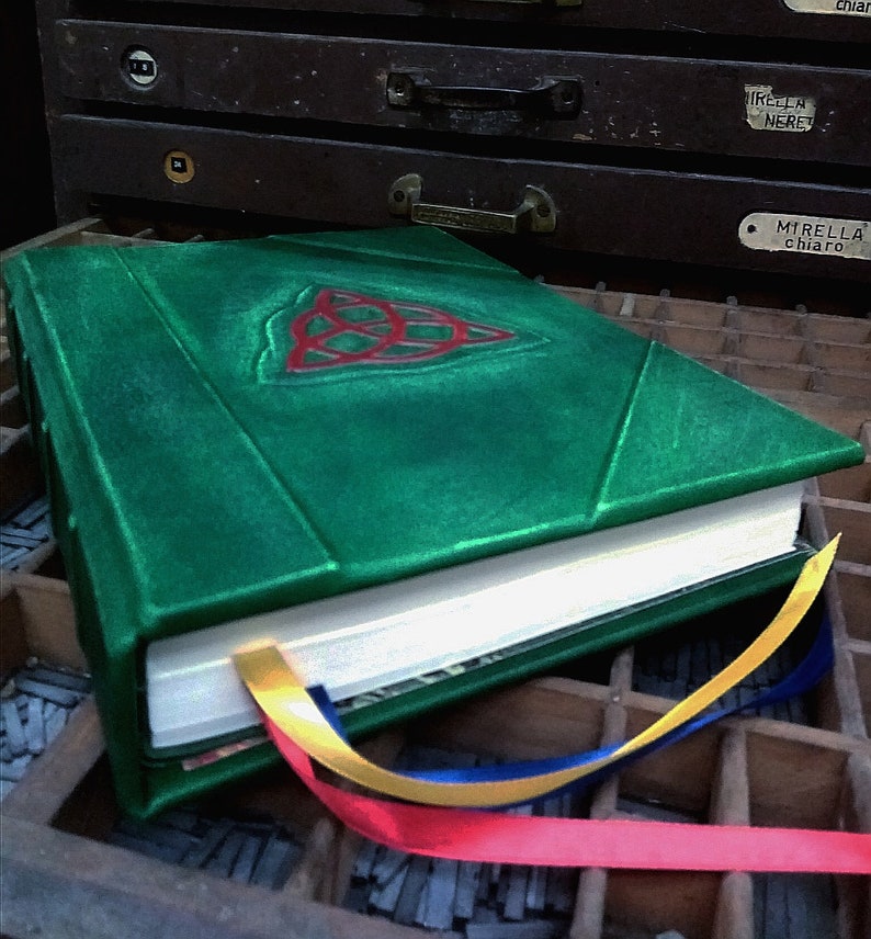 May include: A green leather-bound journal with a red and green Celtic knot design on the cover. The journal is closed and has a white ribbon bookmark. The journal is sitting on a wooden surface with a variety of metal type.