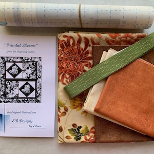 May include: A quilting kit for a "Oriental Blooms" quilt pattern. The kit includes fabric scraps in various shades of brown, orange, and cream with floral patterns, a green ribbon, and a printed instruction booklet.