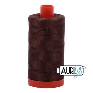 Pode incluir: Uma bobina de fio Aurifil marrom com uma bobina de plástico vermelha.