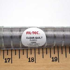 Puede incluir: Una bobina de hilo de color plateado etiquetada "FIL-TEC CLEAR-QUILT CLASS L 12958 LIGHT GREY" en un recipiente de plástico transparente. La bobina se mide con una regla de madera.