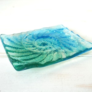 May include: A rectangular glass soap dish featuring a gradient color transition from deep blue to vibrant green. The surface displays a textured, embossed spiral pattern that radiates from the center, with wavy, uneven edges along the perimeter.