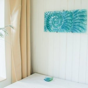 May include: A teal blue glass wall art piece featuring a spiral design. The art is mounted on a white wall with white vertical paneling.