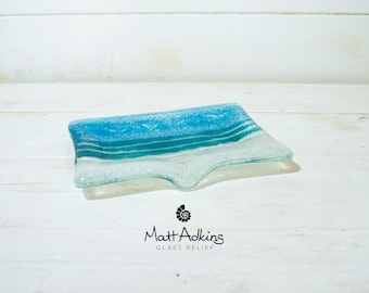 May include: A rectangular fused glass dish featuring a gradient of blue and white tones with a textured, wavy surface. Below the dish, the text reads Matt Adkins GLASS RELIEF with a small spiral shell icon centred above the name.