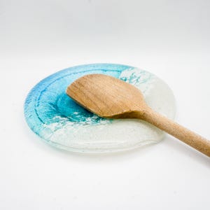 Op de afbeelding: Een blauwe en witte glazen lepelsteun met een golfontwerp. Een houten lepel rust op de lepelsteun.