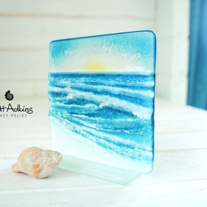 Könnte beinhalten: Eine quadratische Platte aus geschmolzenem Glas mit einer Küstenszene, die eine gelbe Sonne, einen blauen Himmel, weiße Wolken und türkisfarbene Meereswellen zeigt, die auf einen weißen Sandstrand treffen. Der Text Matt Adkins Glass Relief steht links.