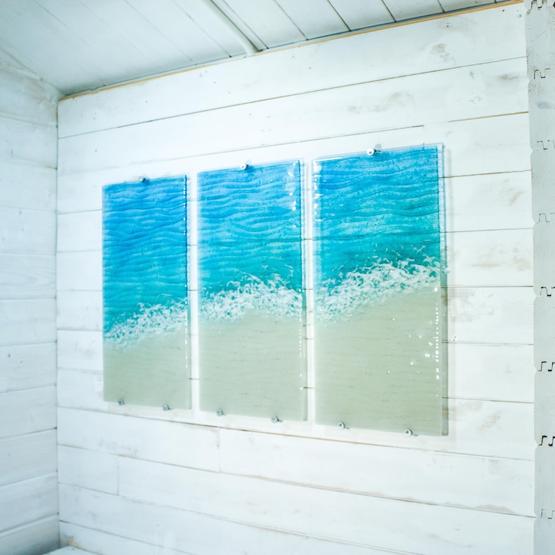May include: A triptych of three rectangular glass wall art panels featuring a textured ocean scene. Each panel displays a gradient from deep blue water at the top, transitioning to turquoise waves with white sea foam, and light beige sand at the base.