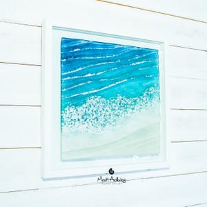 May include: A square glass wall art piece in a white frame depicting an aerial view of an ocean shoreline. The artwork features layers of blue and turquoise glass representing waves with white textured foam meeting a sandy beige beach area.