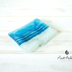May include: A rectangular fused glass soap dish featuring a gradient of deep blue to light blue and clear glass with horizontal white stripes. The bottom right corner includes the text Matt Adkins Glass Relief with a small shell icon.