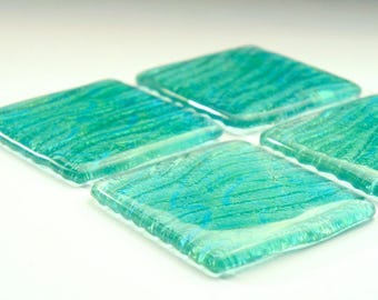 May include: A set of four square glass coasters featuring a translucent teal and turquoise color palette with a textured, wavy pattern embedded within the material. Each coaster has smooth, rounded edges and a glossy, reflective surface finish.