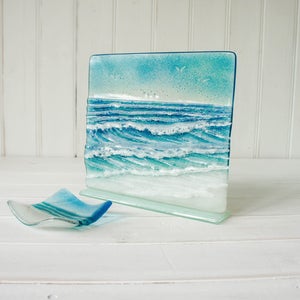 Puede incluir: Un panel rectangular de vidrio fusionado que presenta una escena oceánica texturizada con olas azules de crestas blancas, una orilla arenosa y un cielo azul claro, acompañado de un pequeño plato de vidrio curvo azul y blanco.