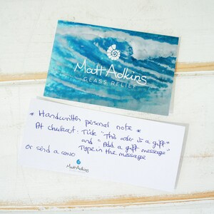 May include: A rectangular business card featuring a blue and white abstract wave design with the white text Matt Adkins GLASS RELIEF and a shell icon, alongside a white card with blue handwritten instructions for adding a gift message.