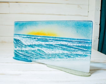 Könnte beinhalten: Ein rechteckiges Kunstobjekt aus geschmolzenem Glas, das eine Küstenszene mit einer hellgelben Sonne über einem strukturierten blauen Ozean mit weißen, brechenden Wellen und einem sandigen Strand zeigt, auf einem klaren Glassockel.