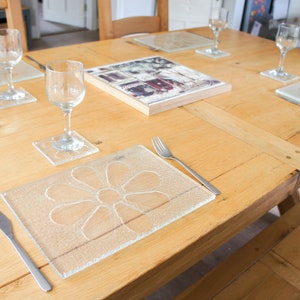 May include: A wooden table set for a meal with four place settings. Each place setting includes a glass, a knife, a fork, and a glass coaster with a flower design. A framed photograph is placed in the centre of the table.
