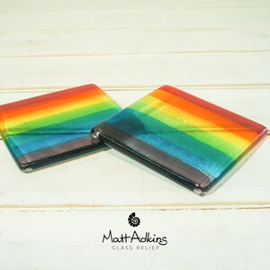 May include: Two square glass coasters featuring a rainbow design. The coasters are made of clear glass with layers of coloured glass to create the rainbow effect. The coasters are on a white wooden surface.