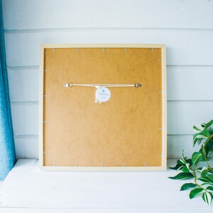 May include: The back of a framed print with a white frame and a brown backing. A white tag with the text "Handmade" is attached to the backing.