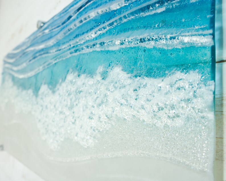 Puede incluir: Primer plano de una pieza de arte de vidrio azul y blanco que representa olas del oc&eacute;ano. La pieza est&aacute; hecha con capas de vidrio, creando un aspecto realista y texturizado.