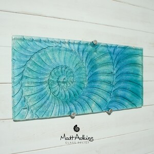 May include: A blue and green glass wall art piece featuring a nautilus shell design. The piece is mounted on a white wall with two silver hooks.