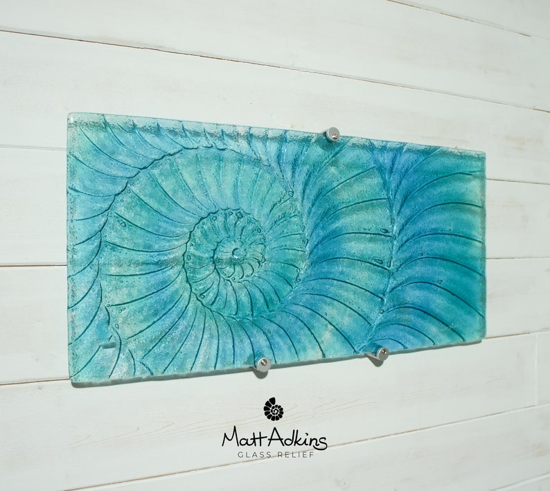 May include: A blue and green glass wall art piece featuring a nautilus shell design. The piece is mounted on a white wall with two silver hooks.