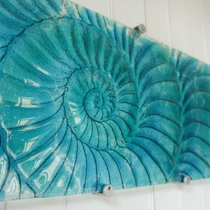 May include: A teal blue glass wall art piece featuring a nautilus shell design. The shell is depicted in a spiral pattern with a light blue and white color gradient.