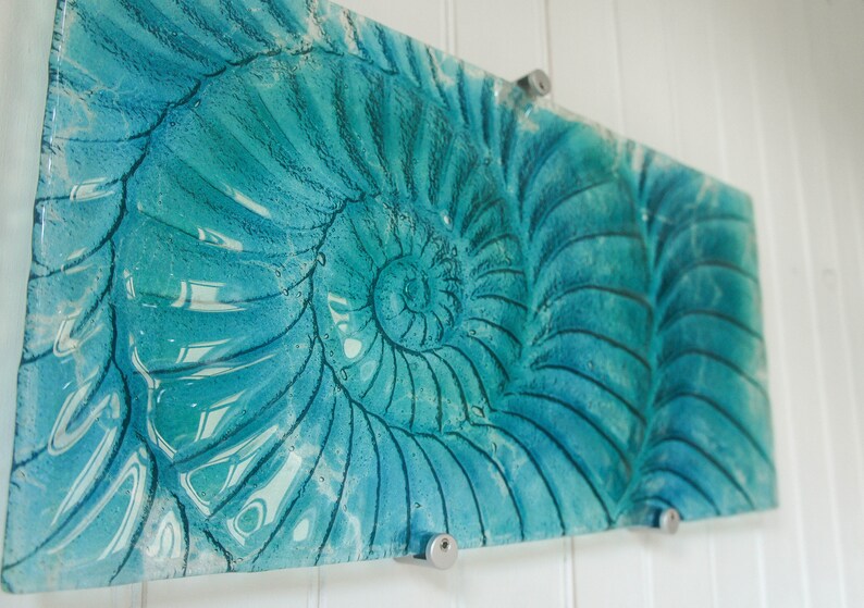 May include: A teal blue glass wall art piece featuring a nautilus shell design. The shell is depicted in a spiral pattern with a light blue and white color gradient.