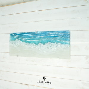 Puede incluir: Una pieza de arte de pared de vidrio que representa una escena de oc&eacute;ano azul con olas blancas espumosas rompiendo en una playa de arena. El arte est&aacute; montado en una pared de madera blanca.