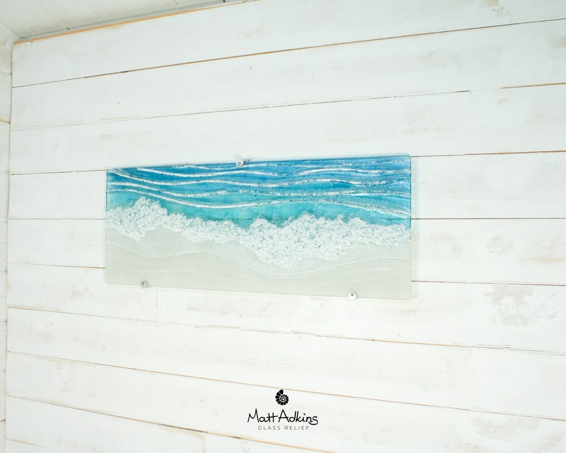 Puede incluir: Una pieza de arte de pared de vidrio que representa una escena de oc&eacute;ano azul con olas blancas espumosas rompiendo en una playa de arena. El arte est&aacute; montado en una pared de madera blanca.