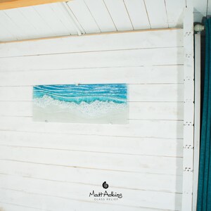Puede incluir: Una pared de madera blanca con una pieza de arte de vidrio azul y blanco que representa olas. La obra de arte se titula "Matt Adkins Glass Relief".